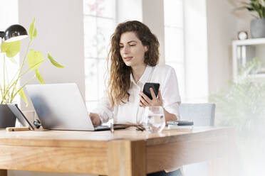 Beautiful female entrepreneur using laptop while holding smart phone at desk in home office - SBOF03600