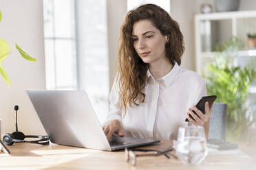 Berufstätige Frau mit Mobiltelefon, die an einem Laptop am Schreibtisch arbeitet - SBOF03602