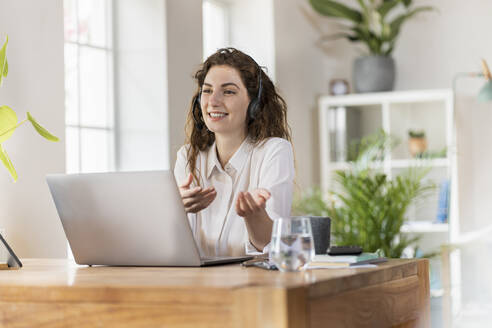 Smiling female woman gesturing while talking through headphones at home office - SBOF03603