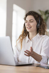 Zufriedene weibliche Kundendienstmitarbeiterin mit Kopfhörern, die einem Kunden am Schreibtisch im Heimbüro etwas erklärt - SBOF03605
