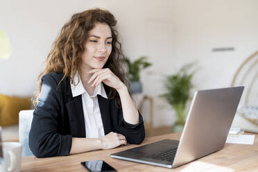 Geschäftsfrau mit Hand am Kinn, die auf einen Laptop im Heimbüro schaut - SBOF03610