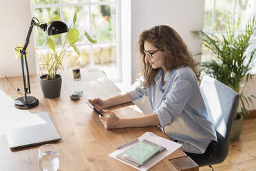 Young businesswoman with eyeglasses looking at digital tablet in home office - SBOF03614