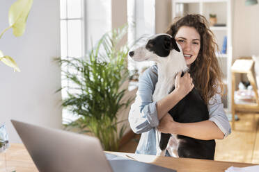 Happy businesswoman cuddling dog at desk in home office - SBOF03619