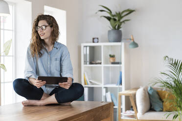 Beautiful smiling woman with digital tablet sitting cross legged on table at home - SBOF03631