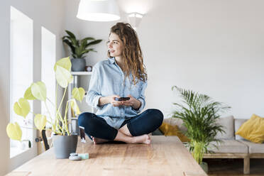 Lächelnde Frau mit braunen Haaren, die wegschaut, während sie ein Handy auf einem Tisch zu Hause hält - SBOF03634