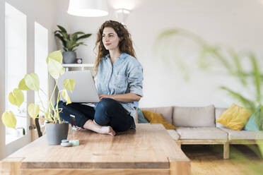 Contented woman with laptop day dreaming while sitting on table at home - SBOF03635