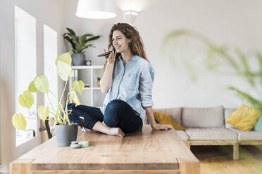 Smiling woman with crossed legs talking on mobile phone at home - SBOF03637