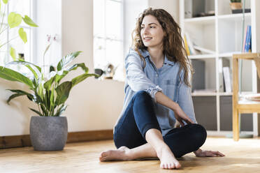 Smiling woman with long hair looking away while sitting on floor in living room - SBOF03640