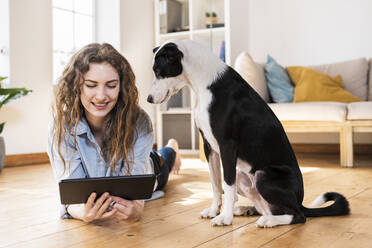 Smiling woman looking at digital tablet while lying on hardwood floor by Jack Russell Terrier in living room - SBOF03641