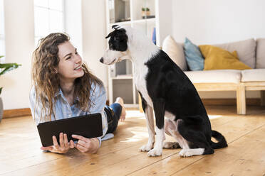 Happy woman with digital tablet looking at dog while lying on floor in living room - SBOF03642