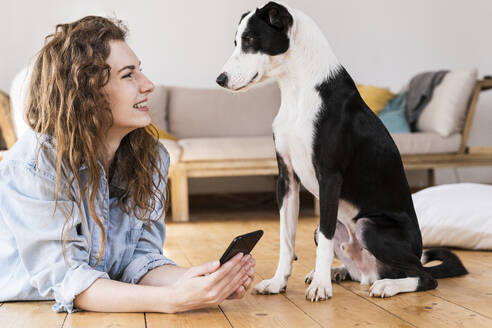 Lächelnde Frau mit Mobiltelefon, die einen Jack Russell Terrier betrachtet, während sie im Wohnzimmer auf dem Boden liegt - SBOF03643