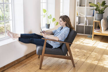 Relaxed young woman with digital tablet sitting with feet up on chair at window in living room - SBOF03648