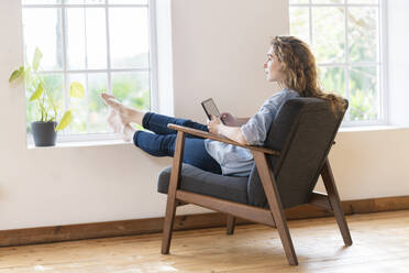 Young woman holding digital tablet while sitting on armchair at home - SBOF03649