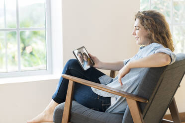 Smiling relaxed woman talking on video call through digital tablet while sitting on armchair in living room - SBOF03651