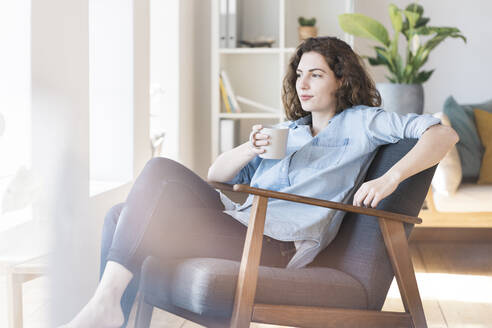 Nachdenkliche Frau, die auf einem Sessel im Wohnzimmer sitzt und Kaffee trinkt - SBOF03652