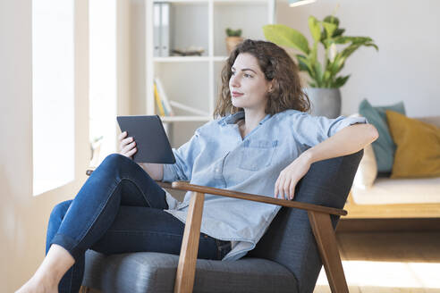 Junge Frau, die auf einem Sessel zu Hause ein digitales Tablet hält und träumt - SBOF03653