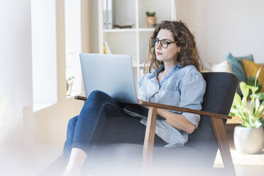 Frau, die einen Laptop benutzt und auf einem Sessel im Wohnzimmer sitzt - SBOF03656