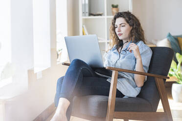 Thoughtful young woman holding eyeglasses while looking at laptop on chair at home - SBOF03657