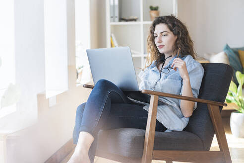 Nachdenkliche junge Frau hält eine Brille, während sie auf einem Stuhl zu Hause auf ihren Laptop schaut - SBOF03657