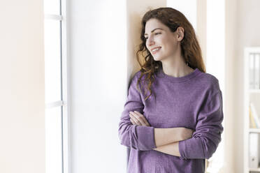 Happy beautiful woman with arms crossed looking through window at home - SBOF03658