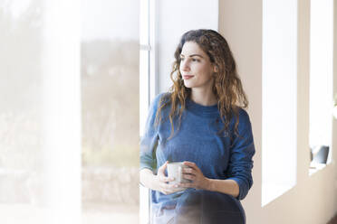 Young woman holding coffee mug while sitting at window in living room - SBOF03660