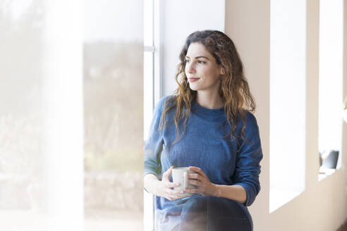 Junge Frau hält Kaffeetasse, während sie am Fenster im Wohnzimmer sitzt - SBOF03660