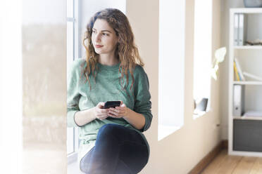 Beautiful young woman day dreaming while holding smart phone at window in living room - SBOF03661