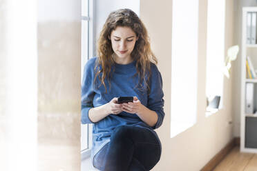 Frau mit braunem Haar, die eine SMS über ihr Smartphone schreibt, während sie am Fenster im Wohnzimmer sitzt - SBOF03662