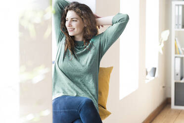 Relaxed woman with hands behind head smiling while sitting at window in living room - SBOF03664