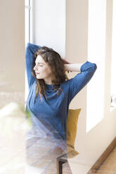 Junge Frau mit geschlossenen Augen entspannt am Fenster im Wohnzimmer - SBOF03665