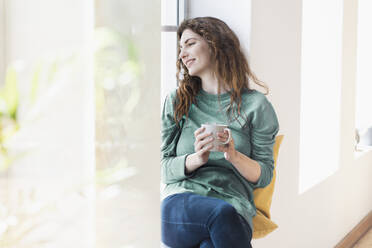 Smiling young woman with coffee mug contemplating while sitting at window in living room - SBOF03666