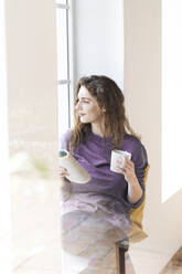 Frau träumt, während sie eine Kaffeetasse und ein Buch am Fenster hält - SBOF03668