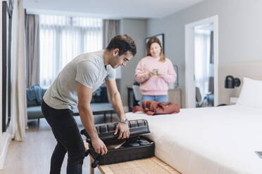 Mid adult man unpacking luggage on bed with woman in background at hotel - DGOF02046