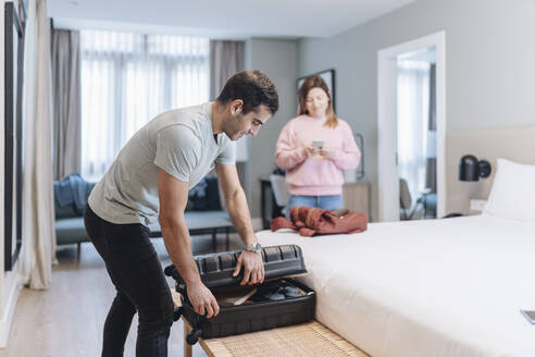 Mid erwachsenen Mann Auspacken von Gepäck auf dem Bett mit Frau im Hintergrund im Hotel - DGOF02046
