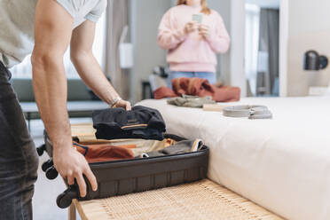 Man removing clothes from suitcase with woman in background at hotel - DGOF02047