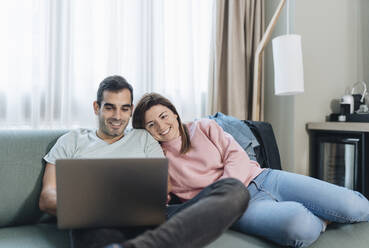 Woman leaning on man using laptop while sitting on sofa in hotel - DGOF02049