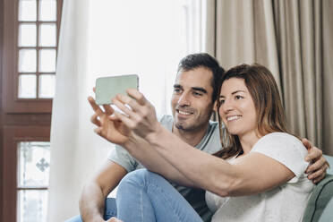 Smiling mid adult couple taking selfie through mobile phone while sitting in hotel room - DGOF02059