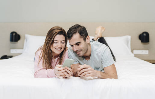 Mid adult couple using mobile phone while lying on bed in hotel - DGOF02064