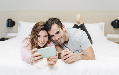 Lächelnde Frau nimmt Selfie mit Mann durch Handy im Hotelzimmer - DGOF02065