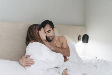 Mann ohne Hemd umarmt Frau, die auf dem Bett im Hotelzimmer sitzt - DGOF02083
