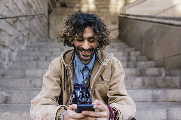 Cheerful man using mobile phone while sitting on steps - XLGF01385