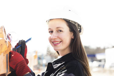 Smiling female engineer writing on clipboard at construction site - SGF02805