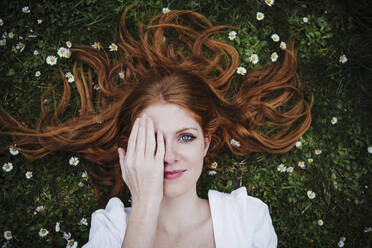 Beautiful young woman covering eye with hand while lying amidst daisy flowers - EBBF02929