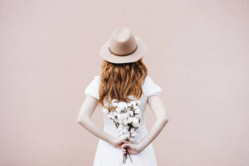 Woman wearing hat holding bunch of cotton plant while standing in front of pink wall - EBBF02937