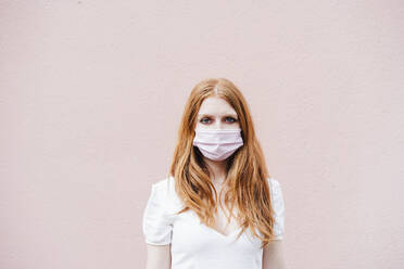 Redhead woman wearing protective face mask while standing in front of pink wall during COVID-19 - EBBF02938