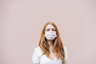 Young woman with eyes closed wearing protective face mask while standing in front of pink wall during COVID-19 - EBBF02939