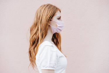 Young redhead woman wearing mask by pink wall during COVID-19 - EBBF02940