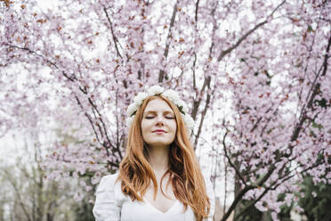 Young woman with eyes closed wearing white flower tiara standing in front of almond tree - EBBF02964