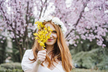 Lächelnde rothaarige Frau, die ihr Auge mit einem Strauß gelber Blumen bedeckt - EBBF02965