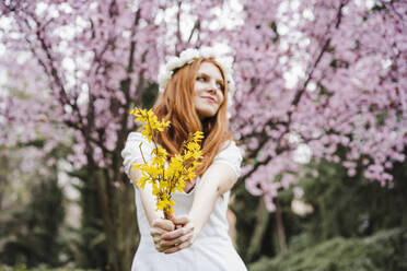 Young woman holding bunch of yellow flowers standing in front of almond tree while looking away - EBBF02968